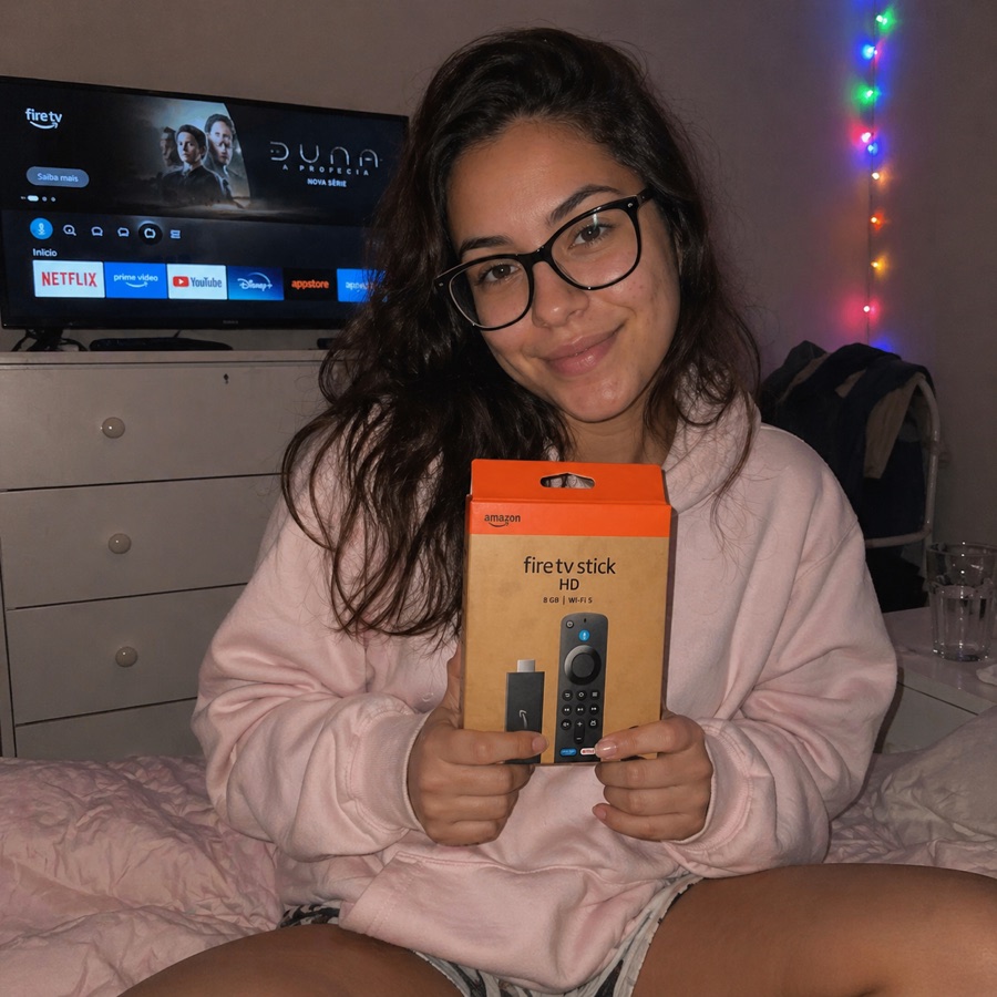 Jovem segurando a caixa do Fire TV Stick HD com TV ligada no quarto